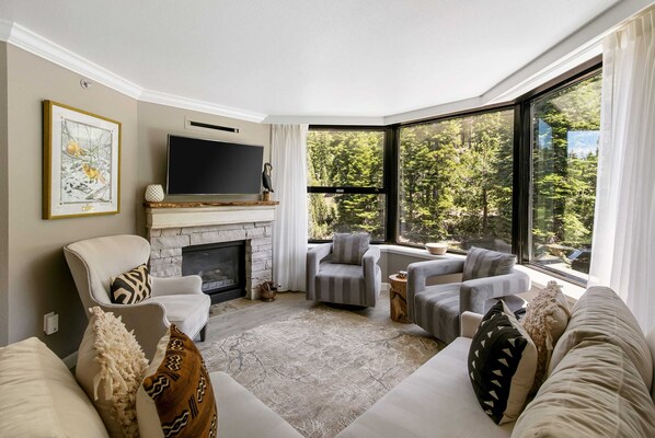 Living Room: L-shaped couch, 3 club chairs, 46” LG TV, gas fireplace, soaring windows framing impressive forested views