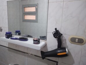 A white bathroom shelf under a wall-mounted mirror holds a hairbrush, blue-lidded jar, and a small tub labeled "Energy." A black hair dryer with orange accents is attached to the wall near a light switch. The wall tiles are white and grey.