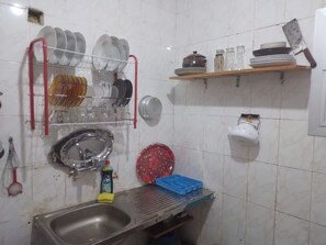 A small kitchen with white tiled walls. There is a sink with dishwashing liquid beside it. Above the sink, dishes are drying on a rack. A red-handled knife is on a wall shelf with additional kitchenware and glasses. An old pot and colander are also visibl