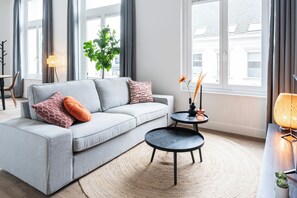 Bright living room with a gray sofa accented by orange and patterned pillows, black nesting tables, natural decor, and large windows for abundant light.