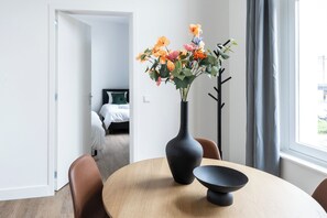 A modern dining arrangement with a light wooden table, sleek leather-look chairs, and a black vase holding a floral centerpiece, set against a bright, open backdrop.