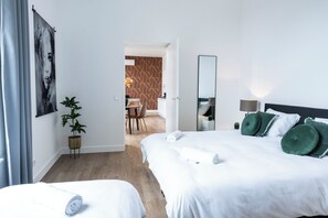 Spacious bedroom with a white bed, green pillows, wall art, a potted plant, and a full-length mirror, leading to a dining area with a bold accent wall.