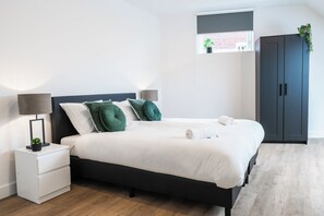 Minimalist bedroom featuring a black-frame bed, crisp white bedding with green decorative pillows, a compact wardrobe, and soft wooden flooring.
