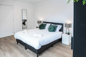 A cozy bedroom with twin beds, dressed in white linens and accented by plush green pillows, flanked by sleek nightstands, modern lamps, and a dark wardrobe.