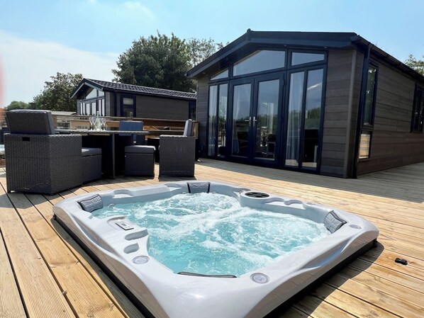 The lodge has entirely glass patio doors that open onto a spacious wooden deck. The deck overlooks a serene outdoor area featuring a sunken hot tub and a four-person dining table. 