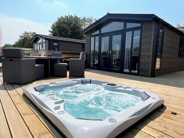 The lodge has entirely glass patio doors that open onto a spacious wooden deck. The deck overlooks a serene outdoor area featuring a sunken hot tub and a four-person dining table.