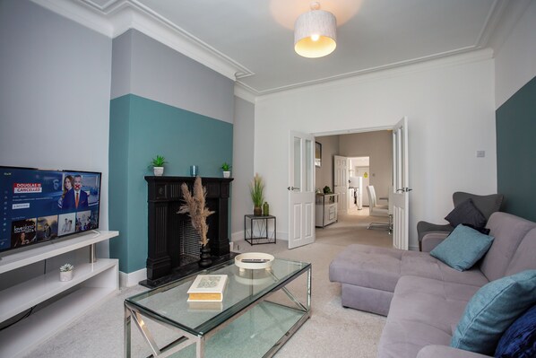 Welcome to our cozy sitting room, featuring comfortable seating for six, a modern flat-screen TV, stylish decor, large windows for natural light, and a coffee table perfect for relaxation.