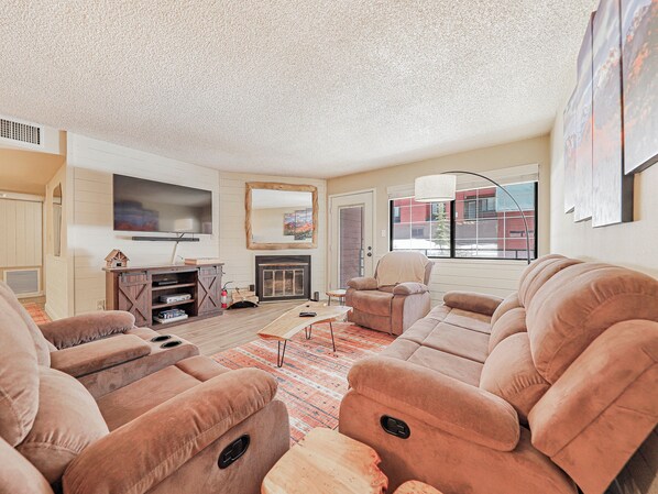Cozy living space featuring a wood-burning fireplace, custom mirror, comfortable sofa and armchair, floor lamp, coffee table, media console, and a 53" smart TV—perfect for relaxing after a day in the mountains