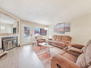 Cozy living space featuring a wood-burning fireplace, custom mirror, comfortable sofa and armchair, floor lamp, coffee table, media console, and a 53" smart TV—perfect for relaxing after a day in the mountains