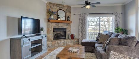 This living room combines comfort and style with its cushy couches adorned with throw pillows, a vintage center table, and a wooden cabinet housing a large TV. The fireplace with elegant accents and the large glass window with floral curtains complete the