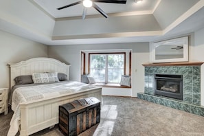 Elegant white wall bedroom featuring a modern fireplace, a white wooden frame wall mirror, and a white-framed bed. The room also includes a wooden ceiling fan and a large window with a cozy sitting area.