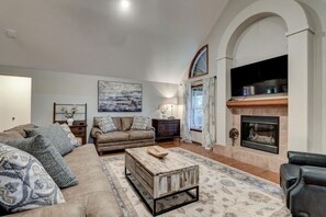 Charming living room with leather couches and throw pillows, vintage wooden cabinets with flower vases, and a contemporary fireplace. The furnishings and accessories include a center table, along with a wall-mounted TV, wall painting, and a window with fl