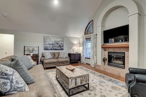 Charming living room with leather couches and throw pillows, vintage wooden cabinets with flower vases, and a contemporary fireplace. The furnishings and accessories include a center table, along with a wall-mounted TV, wall painting, and a window with fl