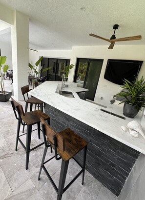 This sleek outdoor kitchen features a modern grill, ample countertop space, a sink, and bar seating, all set against a backdrop of lush greenery for a perfect al fresco dining experience.