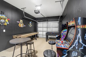 Step into a retro-inspired game room featuring a shuffleboard table, foosball, classic arcade machines, and bar seating. The perfect spot to unwind and challenge friends to a game night!