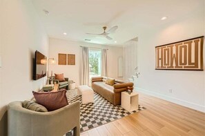 Relax in the bright and airy living room, featuring crisp white walls that create an open, welcoming space. The room is equipped with a flat-screen TV for entertainment, and the wooden-like tile flooring adds warmth and texture, creating a cozy yet m
