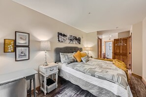 A small bedroom with a large bed, gray tufted headboard, and patterned bedding. The room has a desk, a nightstand with a lamp, framed art on the walls, and a wooden door leading to another room.