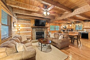 Experience rustic charm in this cozy log cabin living area, featuring exposed wooden beams, a stone fireplace, and plush seating. The open layout includes a fully equipped kitchen and dining space, perfect for relaxing after a day of adventure.