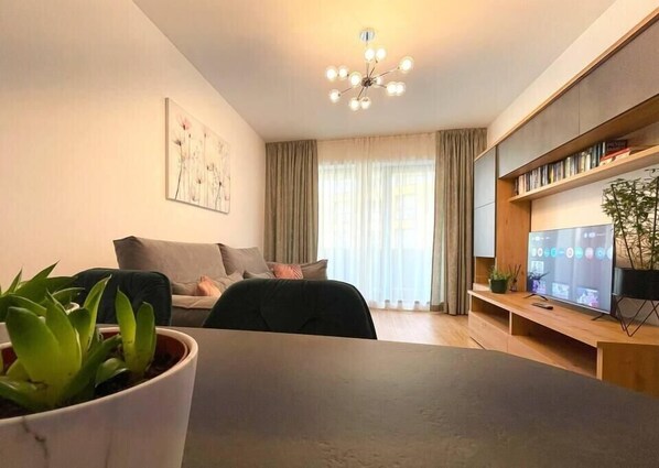 Spacious living room featuring an extensible sofa with decorative pillows, room darkening shades, multi-language library, plants, smart TV with Netfliix, dining table, and free Wi-Fi