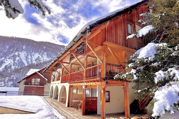 The exterior of the building features a charming wooden facade, with a snow-covered roof and a balcony overlooking the snowy landscape. The building's architecture blends seamlessly with the alpine surroundings, offering a picturesque setting for a peacef