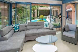 The spacious living area features a plush, grey sectional sofa, ideal for relaxing and enjoying the views.  Large windows offer an unobstructed view of the sparkling pool and surrounding greenery. The modern coffee tables provide ample space for drinks an
