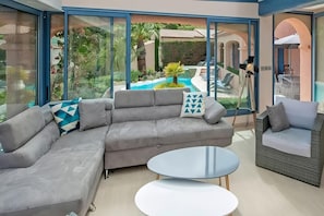 The spacious living area features a plush, grey sectional sofa, ideal for relaxing and enjoying the views. Large windows offer an unobstructed view of the sparkling pool and surrounding greenery. The modern coffee tables provide ample space for drinks an