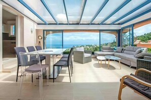The spacious terrace, sheltered by a glass roof, offers stunning panoramic views of the Mediterranean Sea.  The large sectional sofa invites relaxation and conversation, while the dining table with its clear acrylic chairs provides a perfect setting for e
