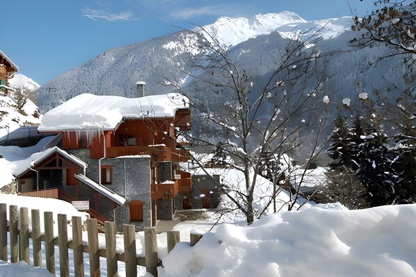 The chalet is nestled in a snowy landscape, with a picturesque view of the snow-capped mountains in the background. The building features a traditional wooden exterior, accented by stonework, creating a charming and cozy atmosphere. The snow-covered roof 