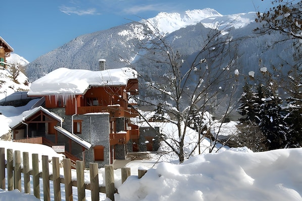 The chalet is nestled in a snowy landscape, with a picturesque view of the snow-capped mountains in the background. The building features a traditional wooden exterior, accented by stonework, creating a charming and cozy atmosphere. The snow-covered roof