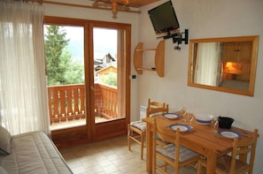 The living area of this chalet features a dining table with four chairs, set for a delightful meal with views of the surrounding mountains. Natural light floods the space through the large French doors, leading to a private balcony. The wooden table and c