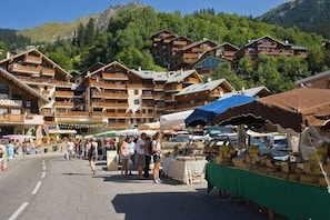 This chalet is nestled within a charming village setting, as evidenced by the vibrant outdoor market taking place just outside. The picturesque mountain backdrop and the bustling atmosphere create a sense of authentic alpine life, offering a glimpse into