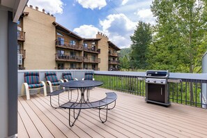 A spacious outdoor deck featuring a round black metal table, four striped chairs, and a gas grill, with an apartment building and trees in the background.