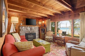 This warm and inviting living room features a stone fireplace, red couch, and large windows overlooking a serene lake. The wooden walls and ceiling beams add to the rustic charm, creating a perfect spot for relaxation.