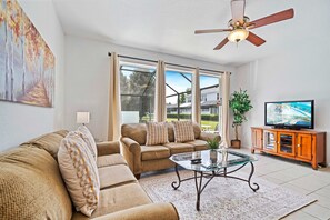 Cozy and bright living area with large windows providing a view of the private pool - Comfortable seating with plush sofas and a table perfect for family gatherings - Ceiling fan and TV for entertainment, creating a relaxing and enjoyable space