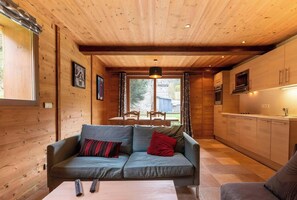 The open-plan living area is a bright and welcoming space with a comfortable sofa, a dining table, and a fully equipped kitchen.  The wood-paneled walls and ceiling create a warm and cozy atmosphere, while the large windows offer stunning views of the sur