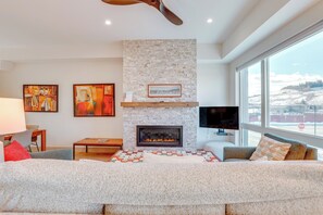 A cozy living room with a stone fireplace, abstract paintings on one wall, a flat-screen TV, and large windows offering an outside view. A ceiling fan is visible above.