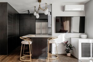 This stylish kitchen island doubles as a breakfast bar with plush gold-accented stools, set beneath a sculptural light fixture and adjacent to a large smart TV for relaxed dining and entertainment