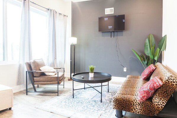 This living room is bursting with personality! A cheetah print sofa takes center stage, complemented by a light brown chair and black coffee table. Plants add a touch of life, and a big window invites natural light. Curl up with a book or movie on the TV. #CozyLiving #ModernElegance