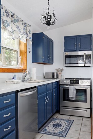Cozy kitchen with blue cabinets, white countertops, and stainless steel appliances. A small window with a curtain adds charm, while a chandelier provides elegant lighting.