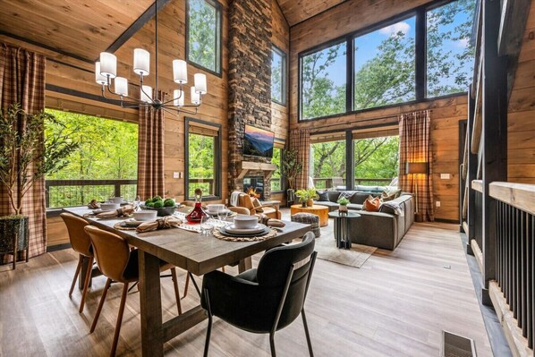 The breathtaking living area of Highland View includes large windows, a floor-to-ceiling stone fireplace, large Smart TV, comfortable furniture, dining table, and kitchen.