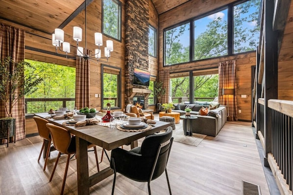 The breathtaking living area of Highland View includes large windows, a floor-to-ceiling stone fireplace, large Smart TV, comfortable furniture, dining table, and kitchen.