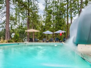 A fenced pool area features a water slide cascading into the shimmering water, surrounded by lounging chairs and colorful umbrellas providing shade and relaxation.