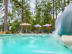 A fenced pool area features a water slide cascading into the shimmering water, surrounded by lounging chairs and colorful umbrellas providing shade and relaxation.