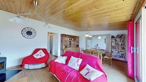 The spacious living room is furnished with a comfortable pink sofa, a matching armchair, and a wooden cabinet. The room features a wood-paneled ceiling and large windows providing plenty of natural light. An open doorway leads to the dining area, while a