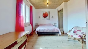Inviting bedroom featuring a comfortable double bed with a patterned duvet. The light and airy space boasts a large window with bright red curtains, a charming rattan chair, and a simple desk. A cozy wood-paneled ceiling and a contemporary poppy painting