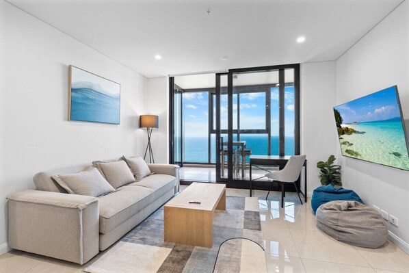 Bright living room filled with natural light from the coastline, featuring a comfortable king sofa bed and a smart TV.