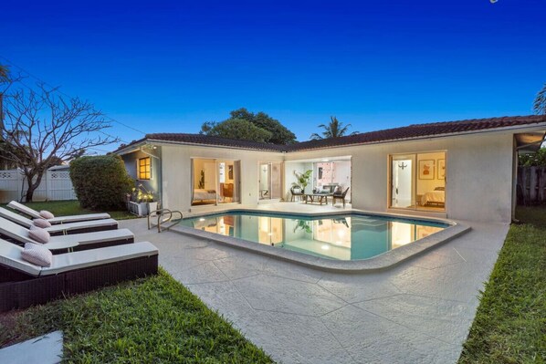 A serene evening view of a modern property with an inviting pool, glowing interiors, and cozy lounge chairs, perfect for relaxation.