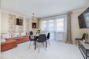Another living room angle with a dining area and a large window that brings in natural light. The table is set with plates, and the warm-toned furniture adds to the welcoming feel of the room.