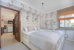A bright bedroom featuring a large bed with soft white bedding and light wood furniture. The room has floral wallpaper and a window allowing natural light to fill the space.