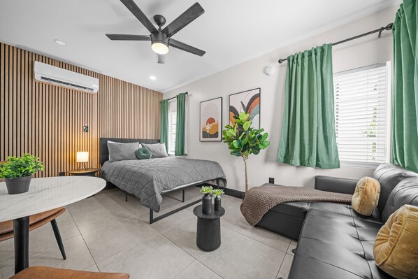 This inviting studio features a cozy queen-size bed, a comfortable leather sofa bed, and stylish decor with vibrant wall art and greenery. Natural light from large windows and a wood-accented wall create a warm, relaxing atmosphere.